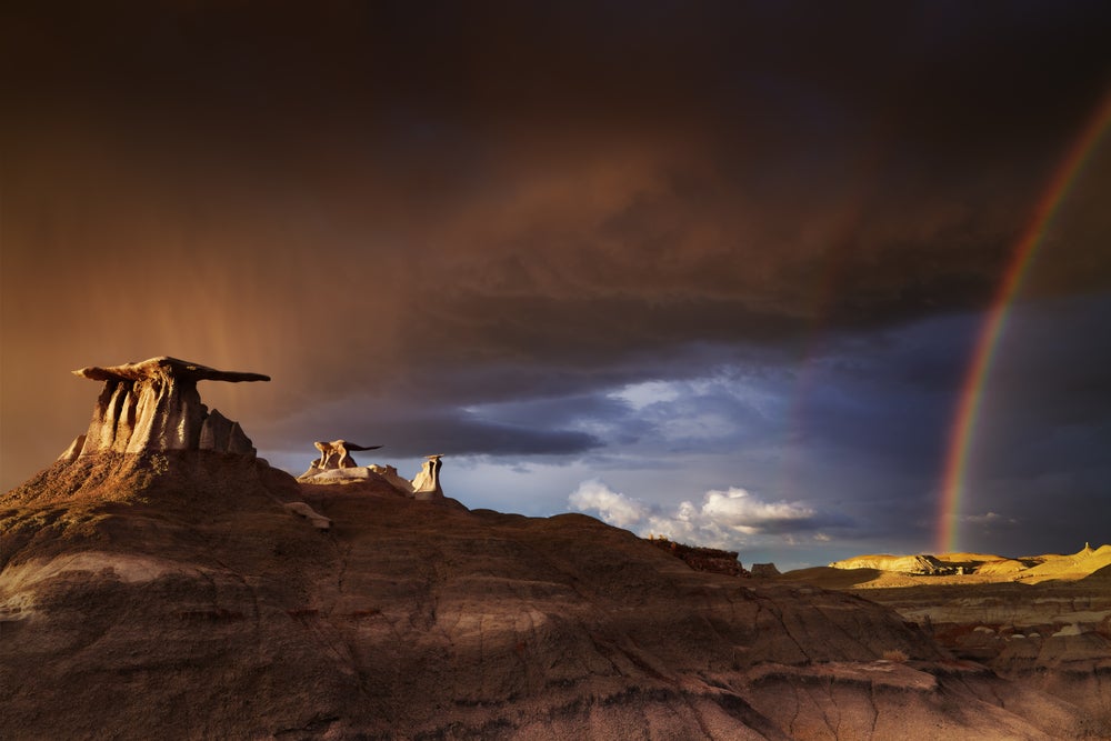 wide angle view of the bisti badlands seen below rolling storm clouds and a vibrant rainbow