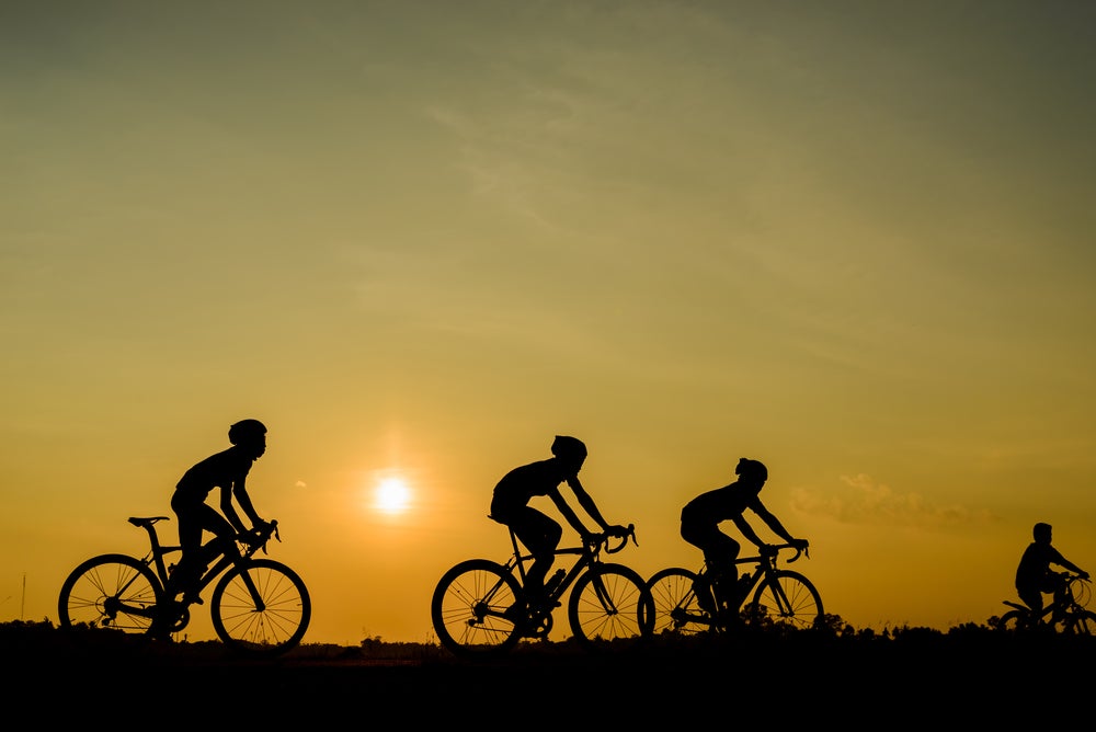 four silhouettes of cyclists ride on a flat plain while the sun sets behind them