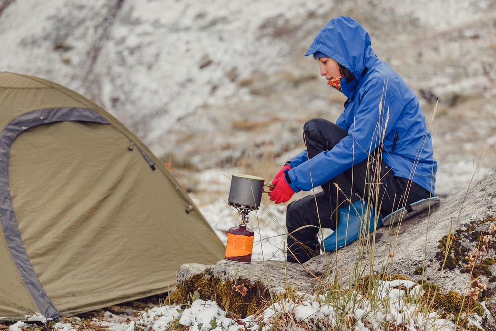 Woman in a blue hooded rain coat holds pot over a gas fire while winter tent camping