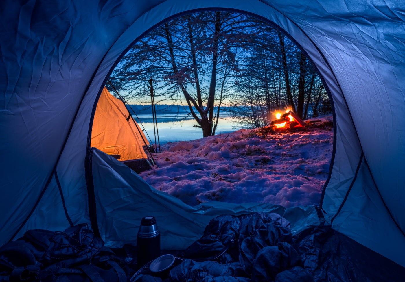 A snowy campsite near a lake with campfire and second tent in the distance as seen from the inside of a tent in the winter