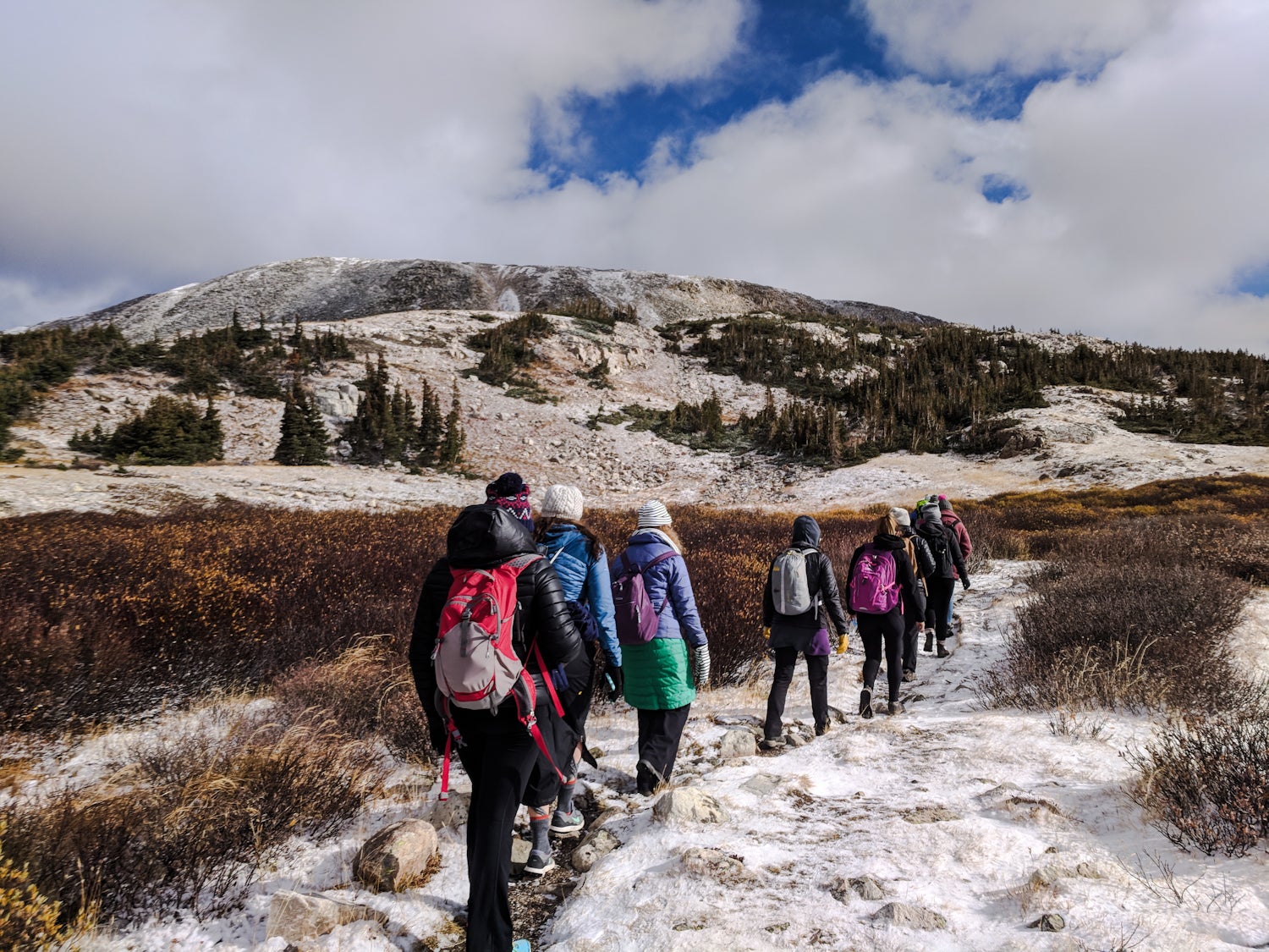 Women hiking while bundled in puffer jackets and beanies with day packs, headed up a trail through snow in Wyoming.