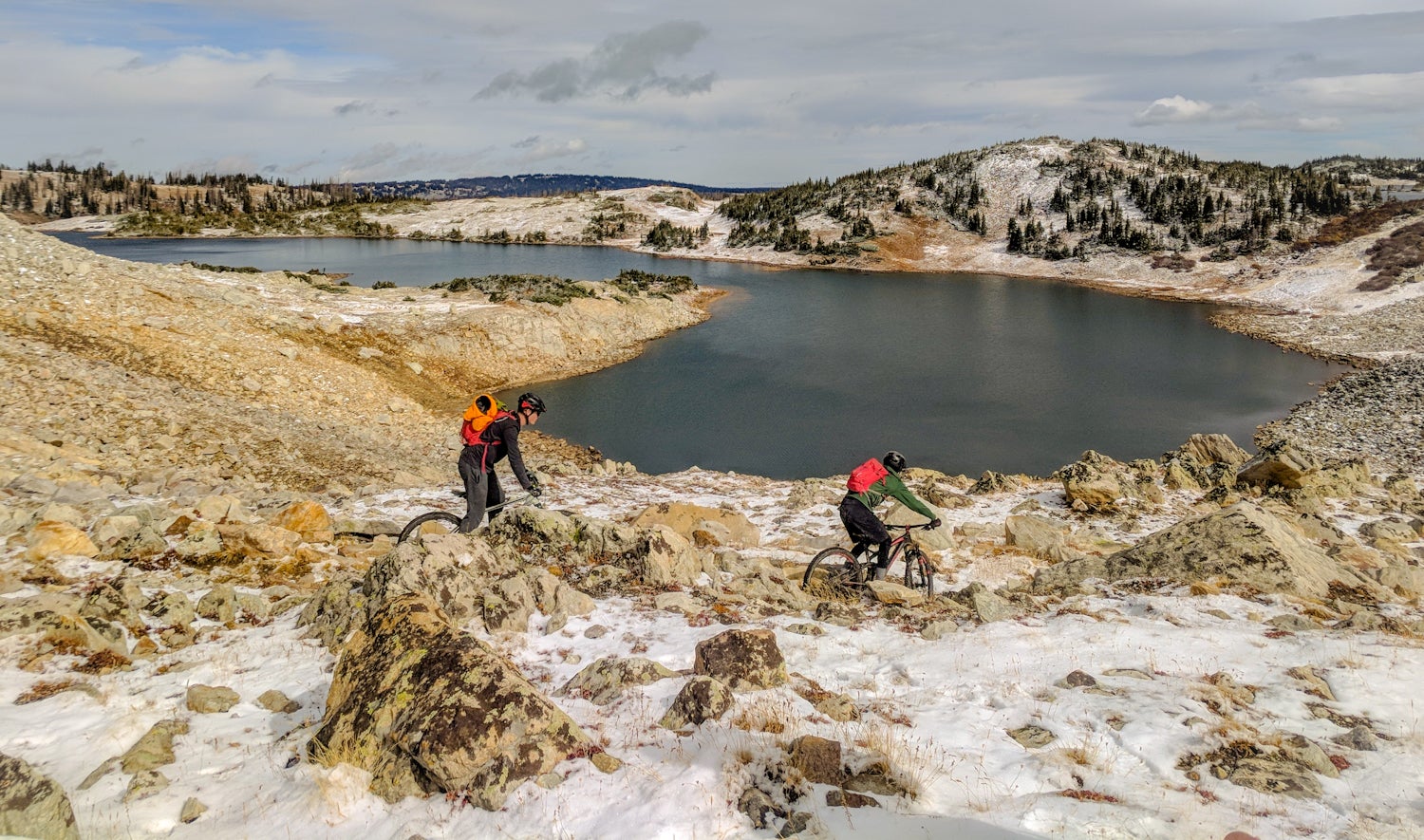 Two mountain bikers manuever through rugged landscape and snow patches beside an alpine lake.
