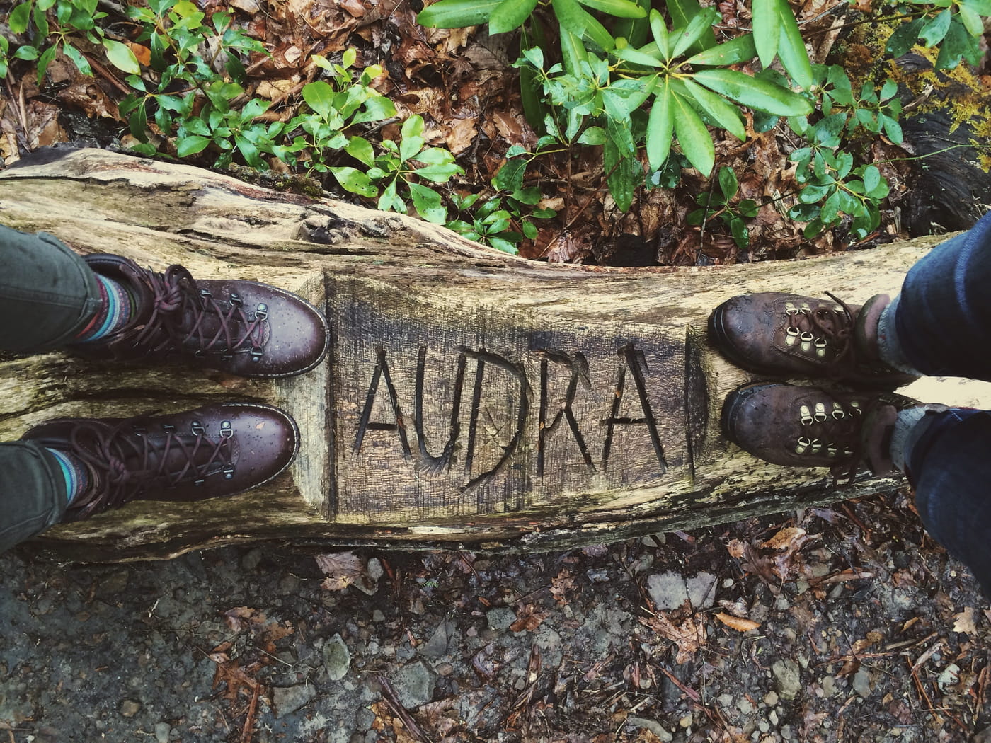 Two people in leather boots standing on a carved log displaying the word Audra.