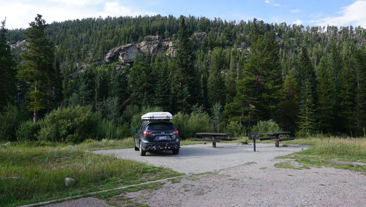 Black SUV with white roofbox parked in drive in campsite with picnic tables in Colorado.