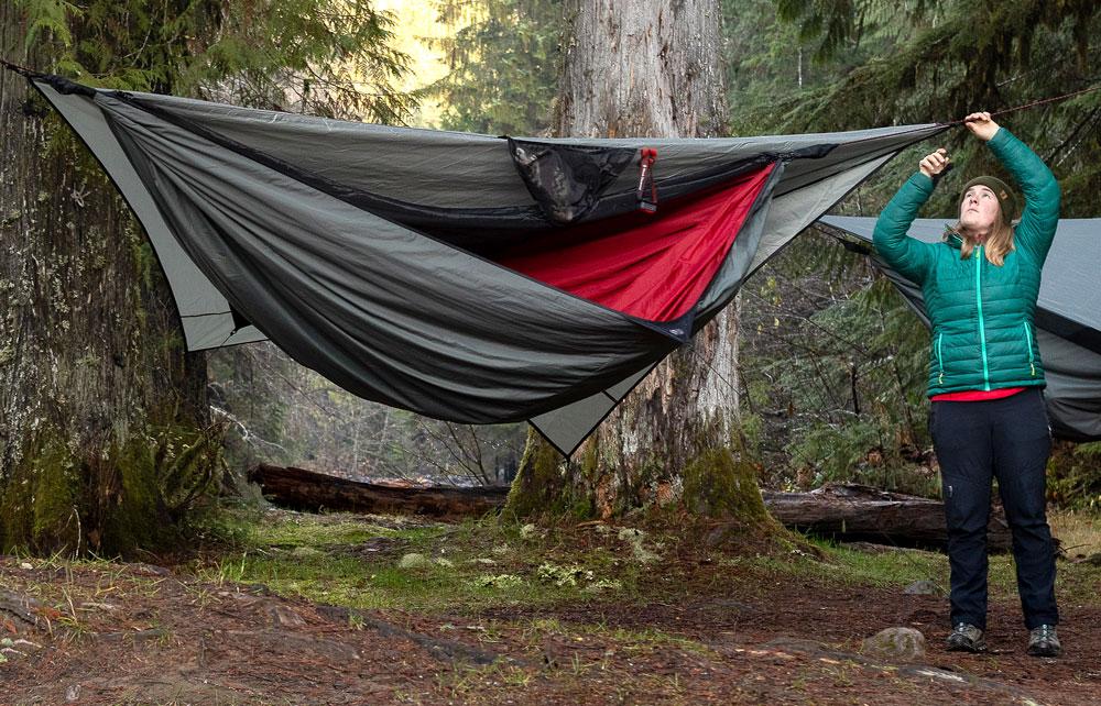 a woman in a blue jacket ties one side of a 4-season hammock to a tree