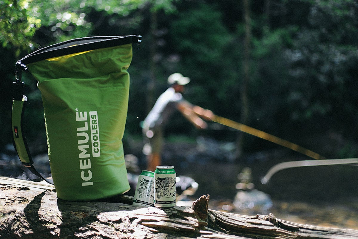 a backpack cooler on a log with two cans of beer, and a man fishing in the background