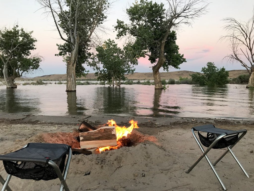 campfire on sandy beach with two camping chairs with trees and water in the background