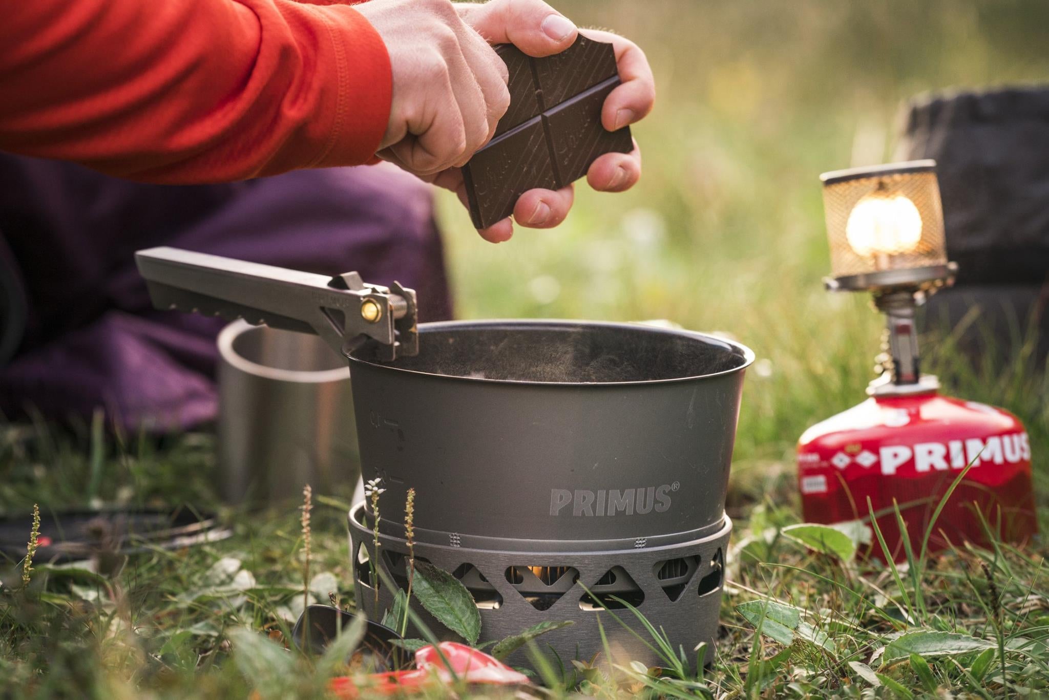 a hand dropping chocolate into a pot over a primus portable stove