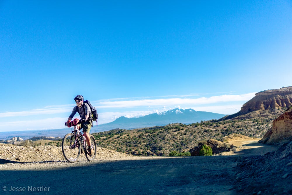 Man biking on trail with mountains and red rock in background