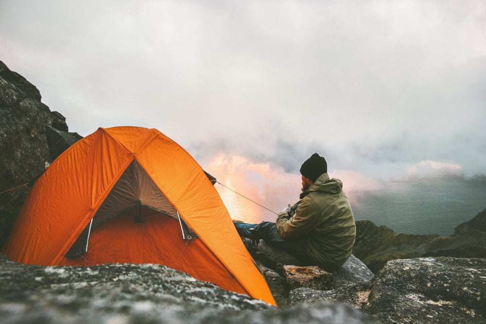 Man sitting on rocky mountainside beside an orange tent while looking out at the sunrise.