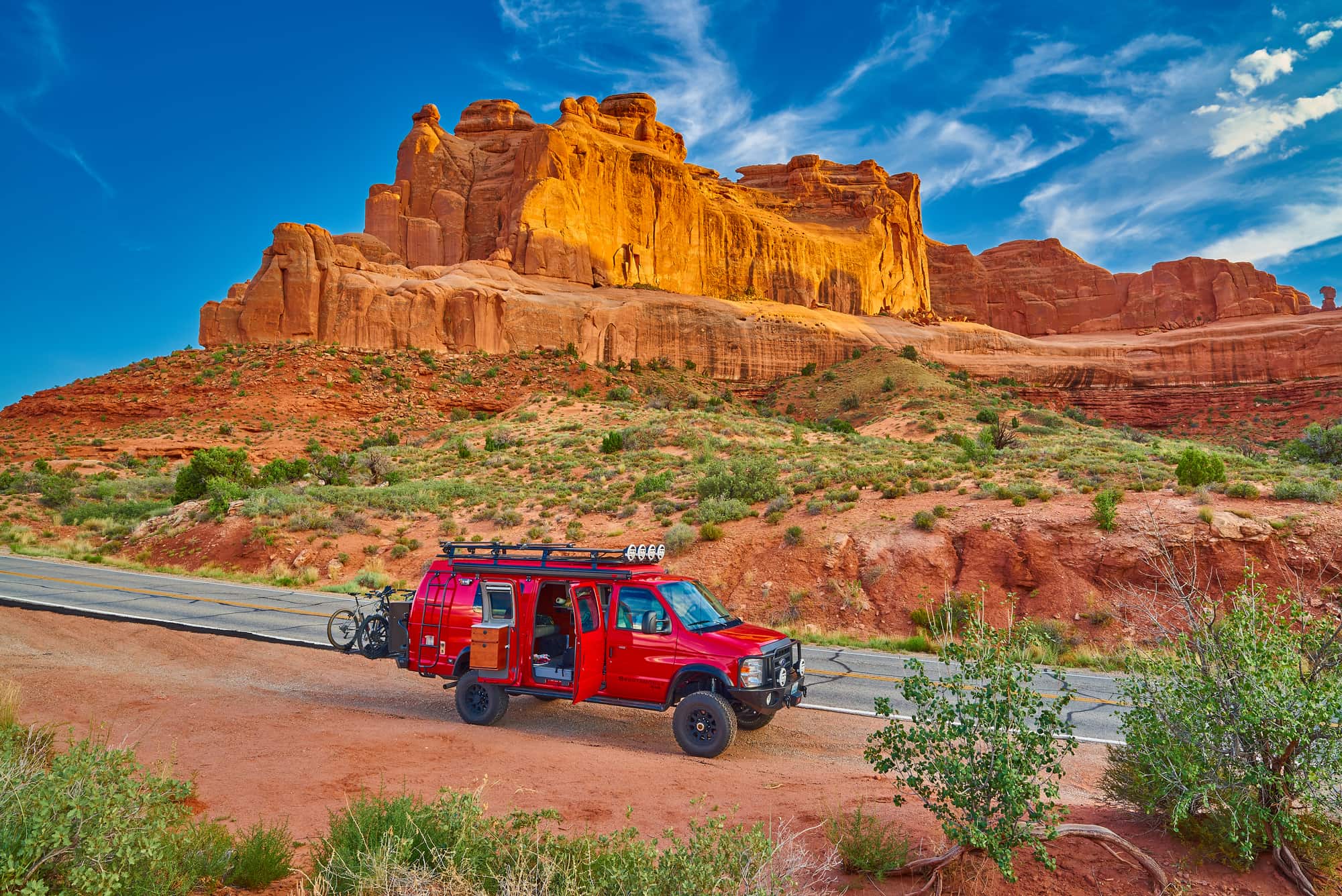 Red off roading adventure van with bikes on the back parked beside red rock formations of Arches Nation Park.