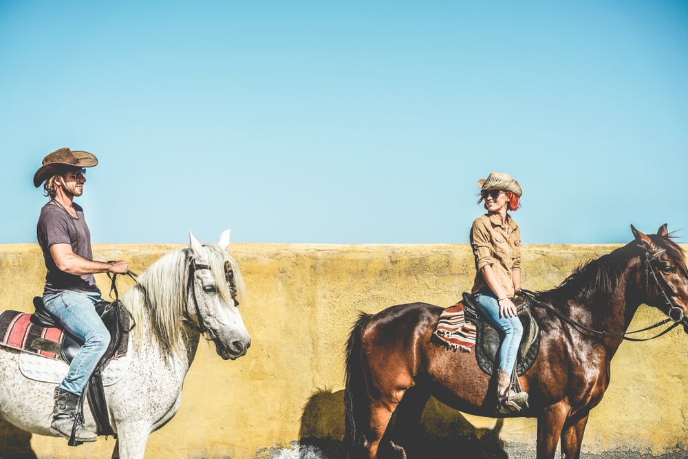 Couple wearing cowboy hats pause on horseback in front of adobe wall