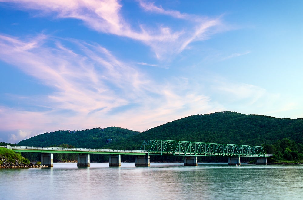 Panoramic view of rolling green hills with green bridge over river. 