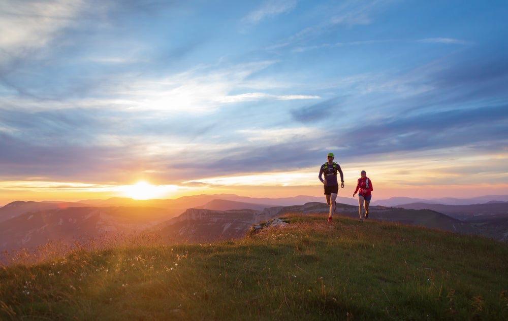 Two trail runners running on a grassy peninsula at sunset, with mountain ranges visible in the distance. 