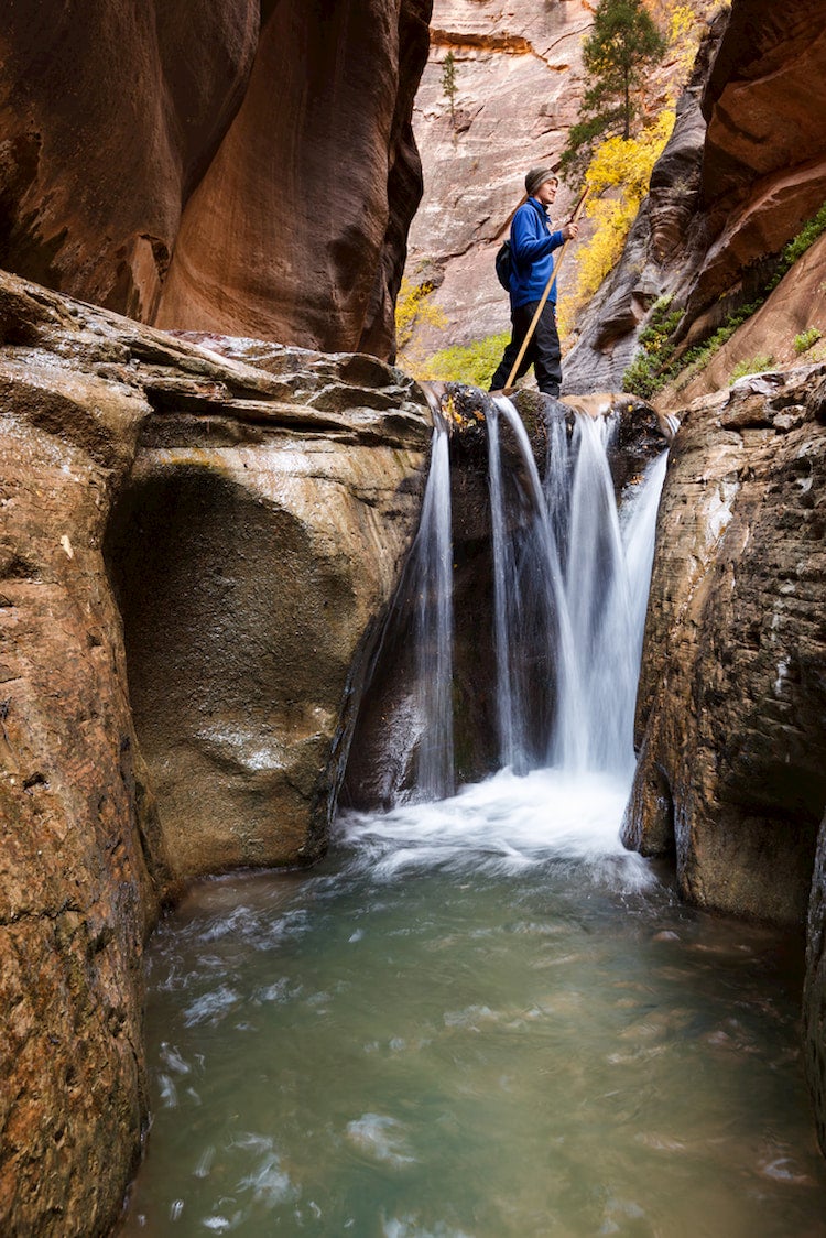 A man standing above a waterfall in orderville slot canyon with a walking stick.