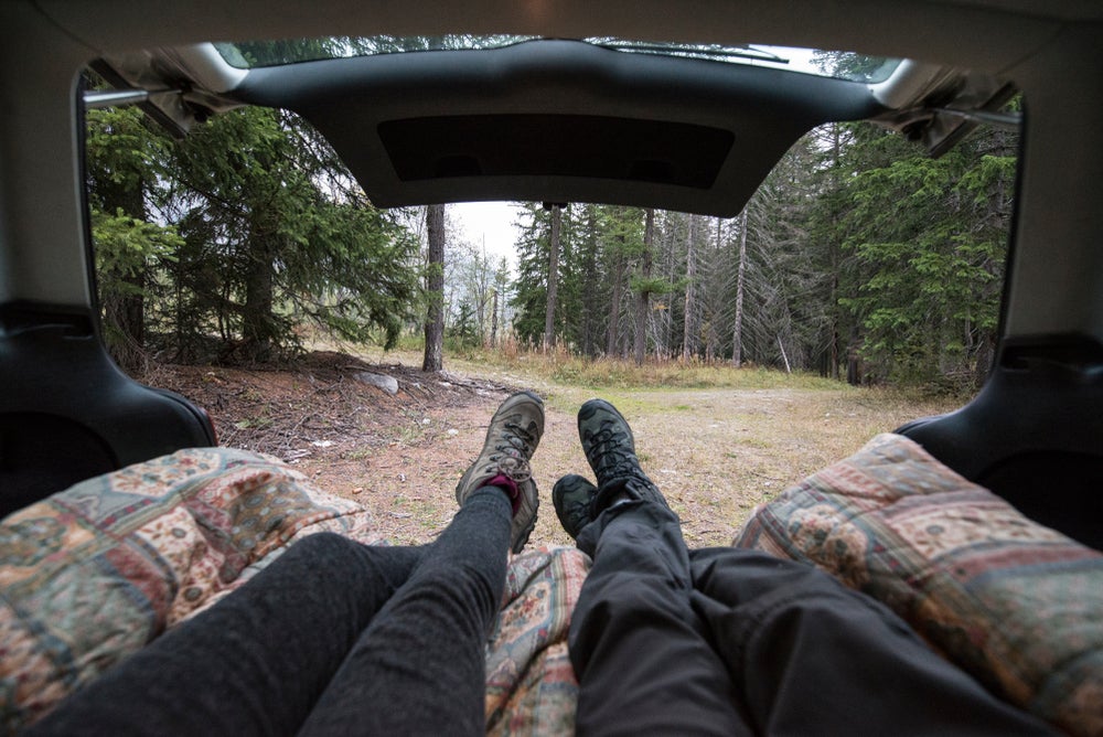 Couple sitting on a mattress in the back of a car in a forest with their feet sticking out.