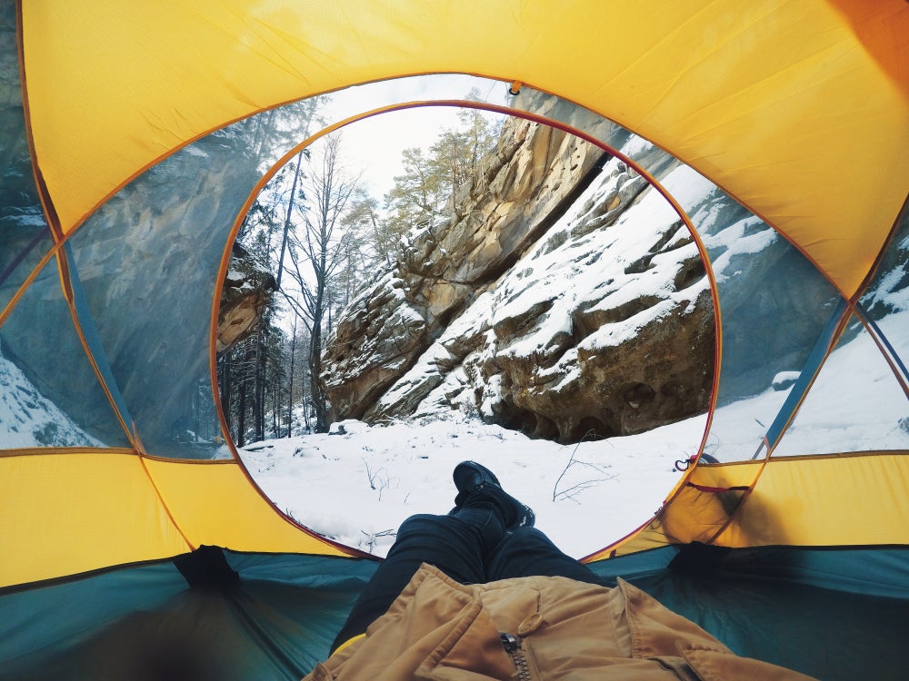 Camper resting on their back sticks feet out into snow through tent's circular opening 