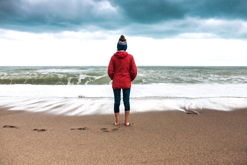 Woman standing in winter clothes while barefoot on the beach, facing the breaking tide.