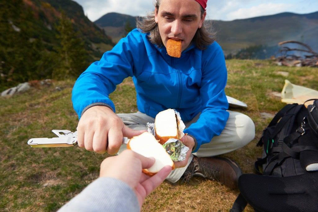 man slicing cheese held by photographer while seated in field