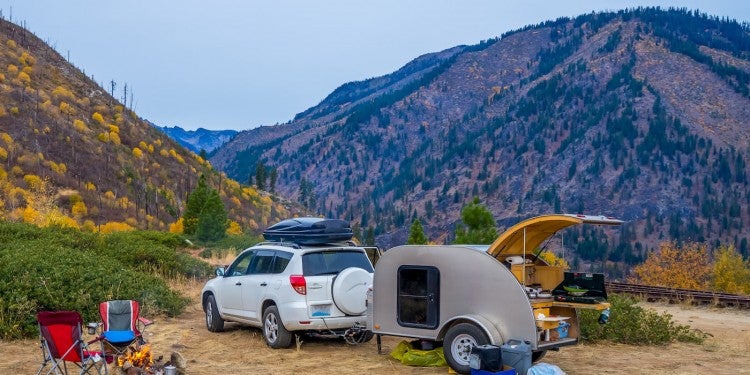Small SUV towing a teardrop trailer parked in the mountains on a dirt road at a campsite.