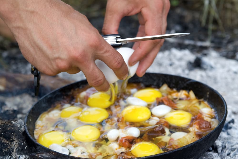 a person cracking eggs in cast iron skillet over campfire coals