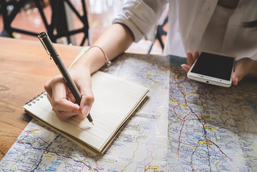 Woman looking at map and writing in notebook with phone in her hand 