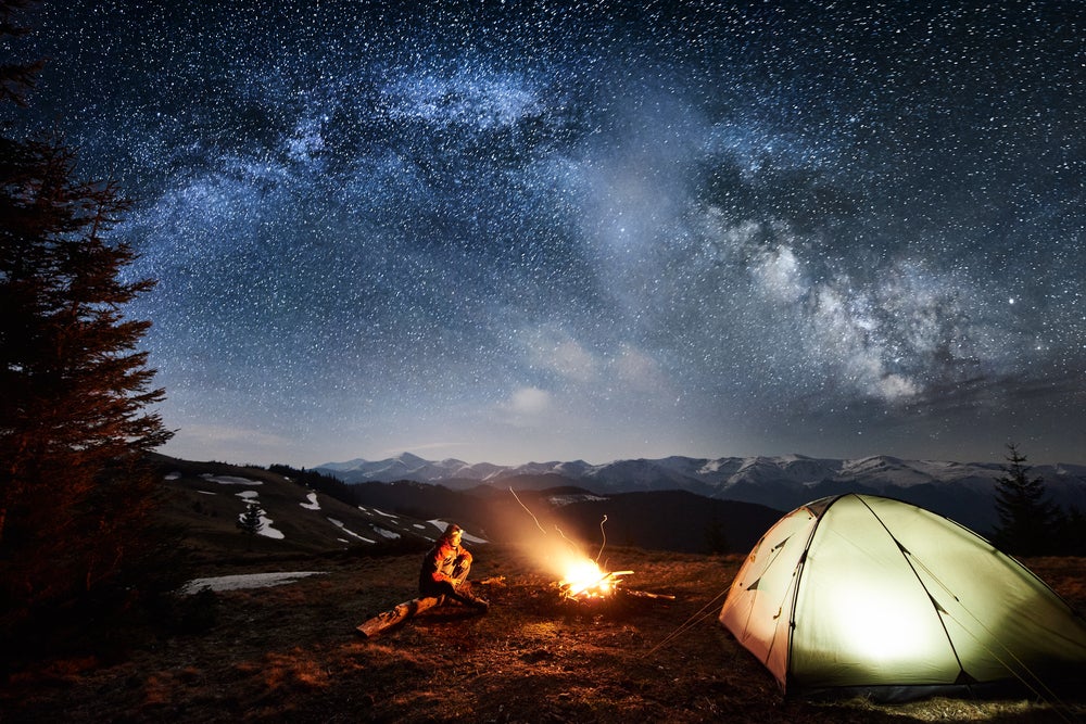 Long exposure of lone camper sitting fireside below stars and beside illuminated tent