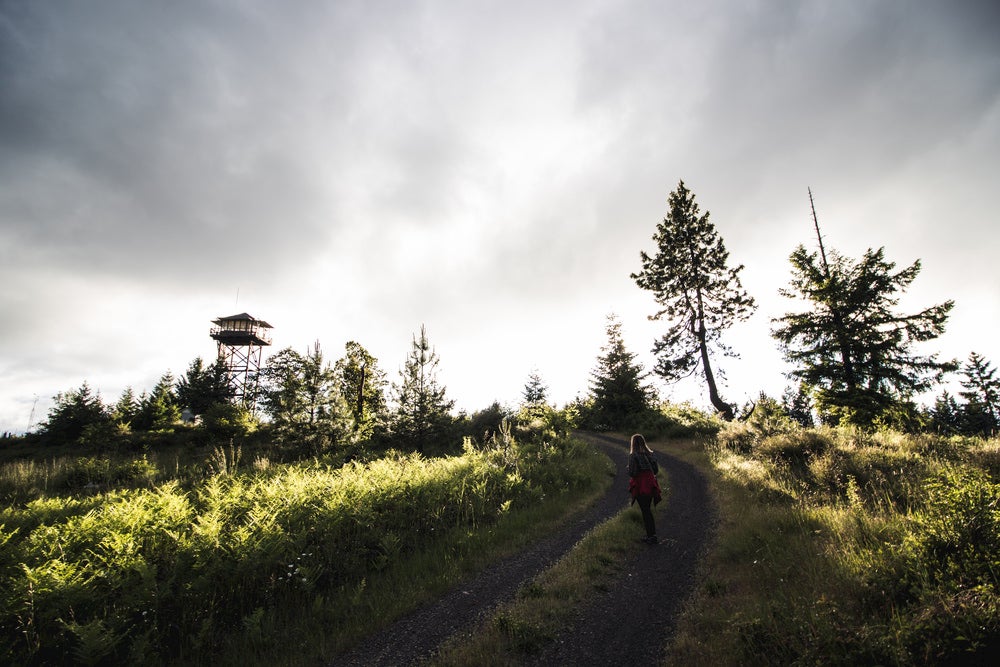 Sun peeks through clouds and trees as woman walks up gravel road to fire tower in the distance