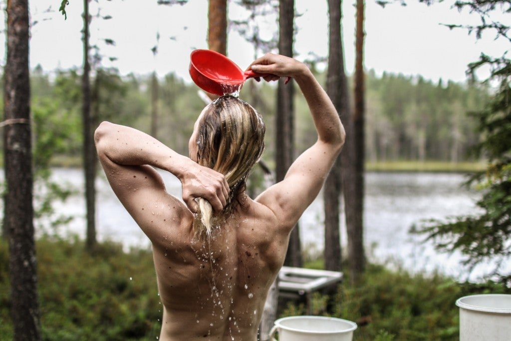Women pours water from a red pot over her head while showering in the woods