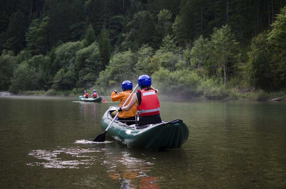 two people in helmets and vests paddling down a river in an inflatable canoe.