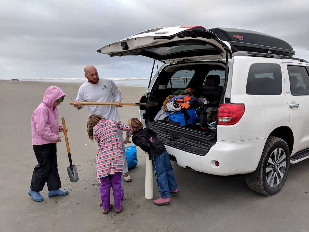 three children look into a car trunk while a man puts a shovel inside