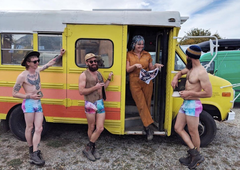 three men in bathing suits surround a woman in converted school bus as she holds a custom bathing suit