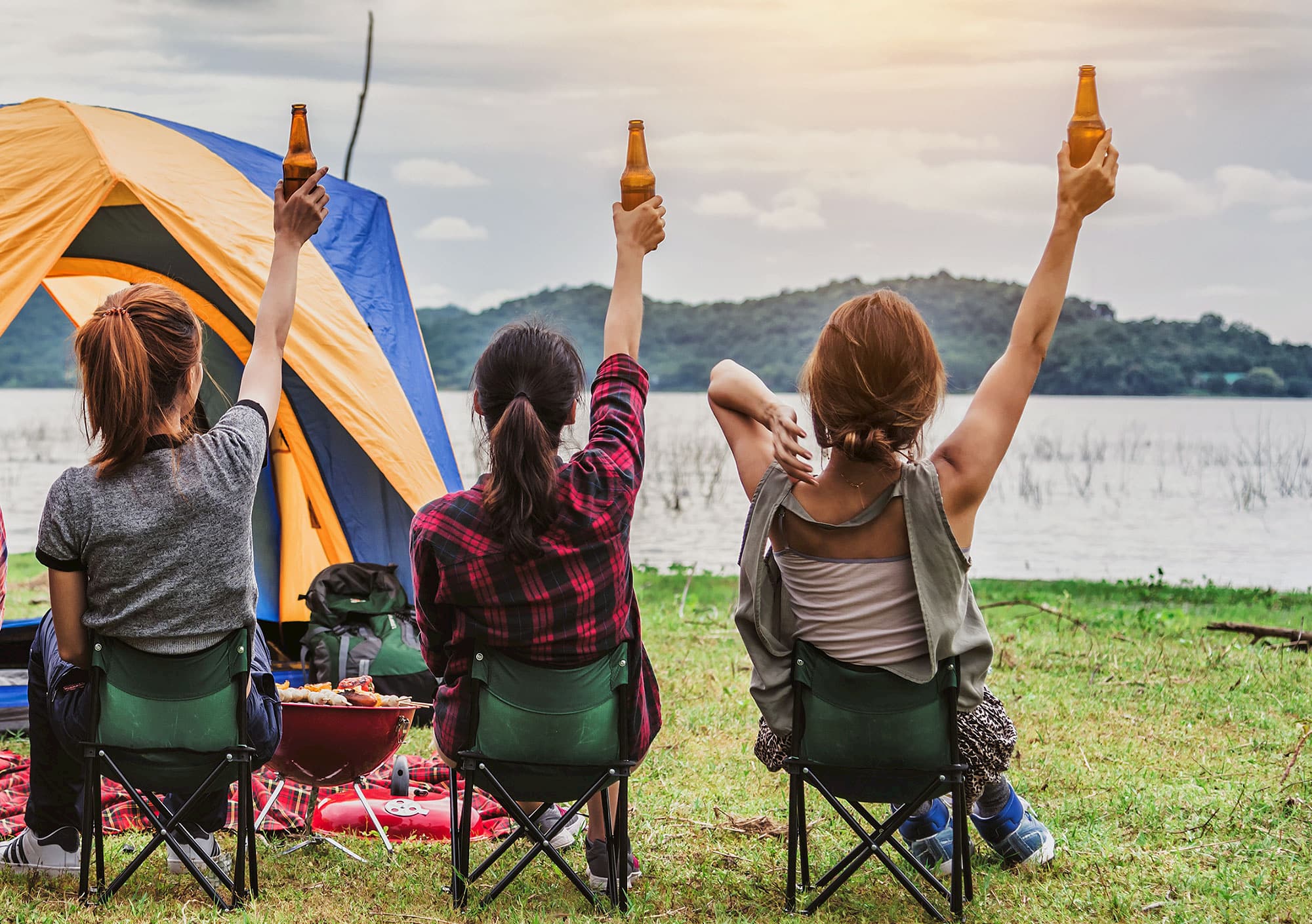 three women holding beers near a tent in chairs while camping