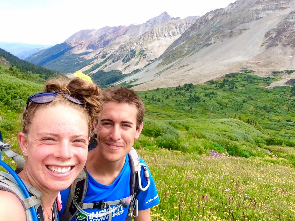 Two people hiking with wildflowers and mountains in background 