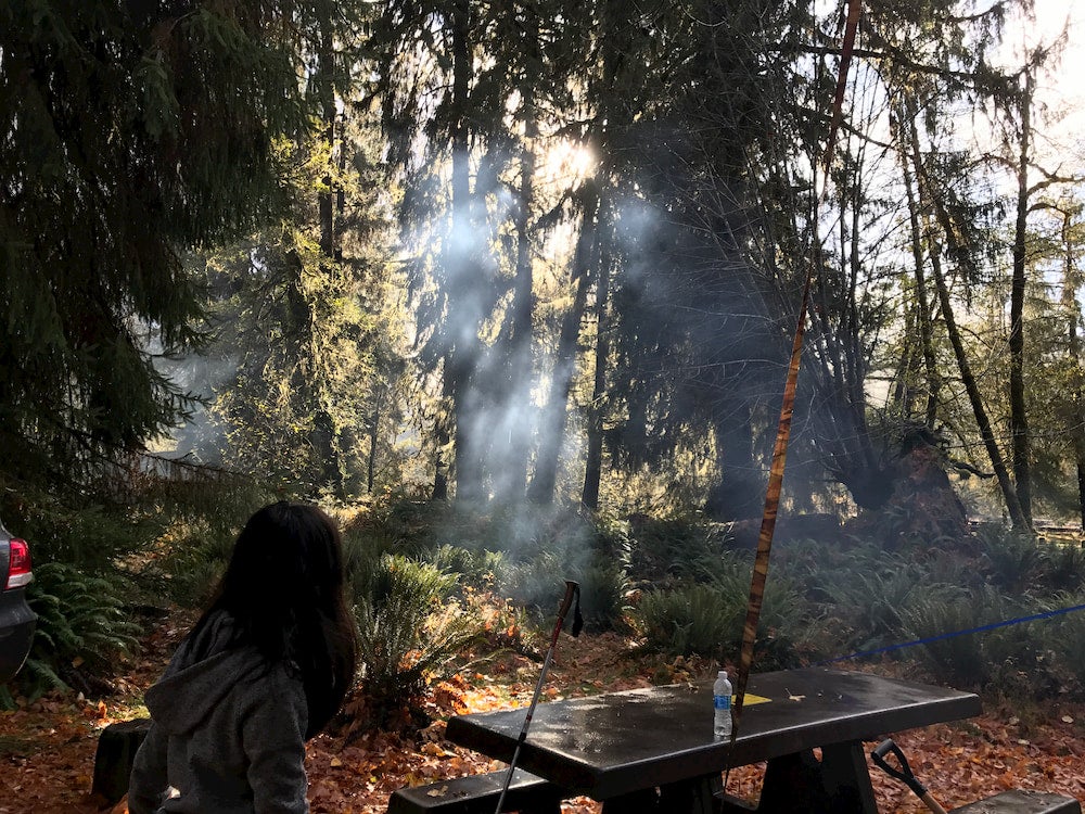 Girl sitting at picnic table in forest with light shining through trees and campfire smoke