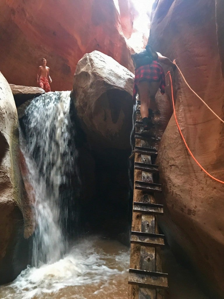 a woman climbing a wood ladder to reach the top of a small waterfall in a slot canyon in utah