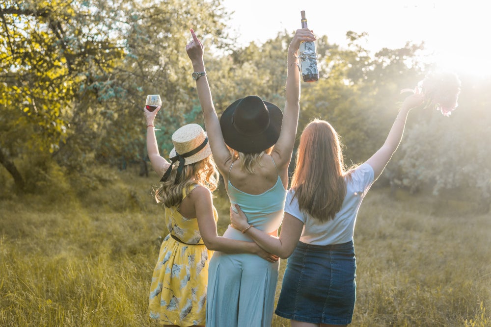 Three women holding drinks and flowers with trees surrounding them at a camping bachelorette party