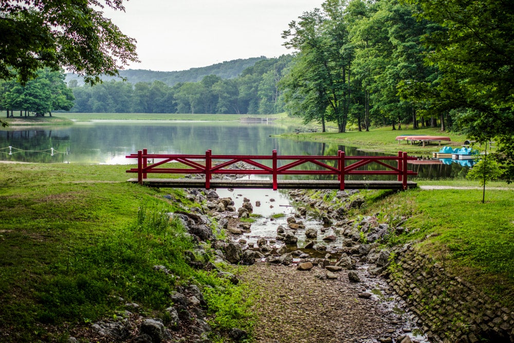 Small red footbridge stretches over rocky riverbed leading away from lush green lake.