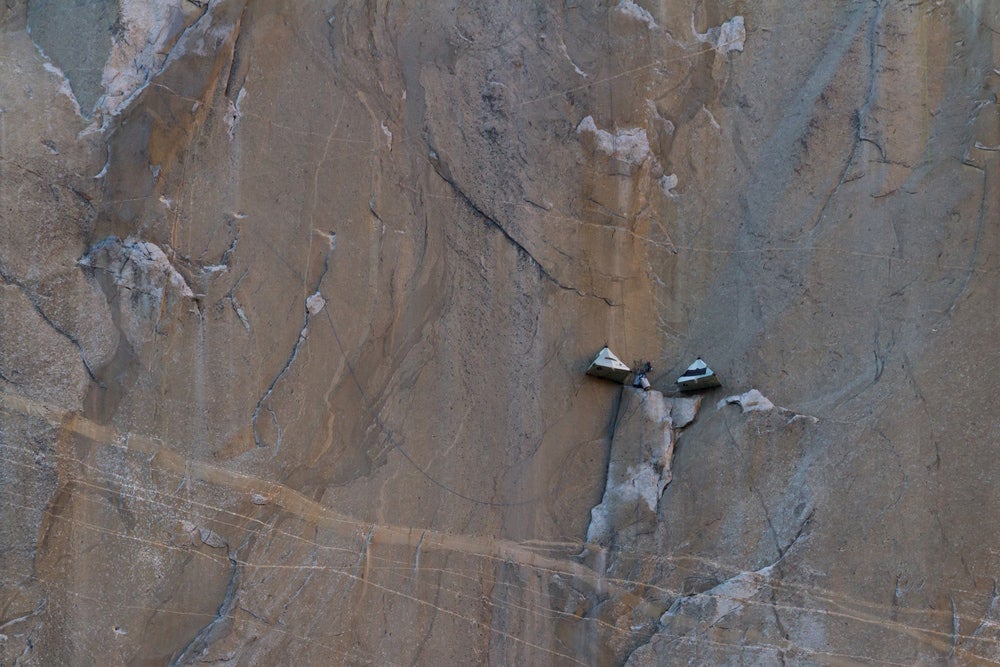 Two vertical campers hanging on the side of a large rock face, seen from a distance.