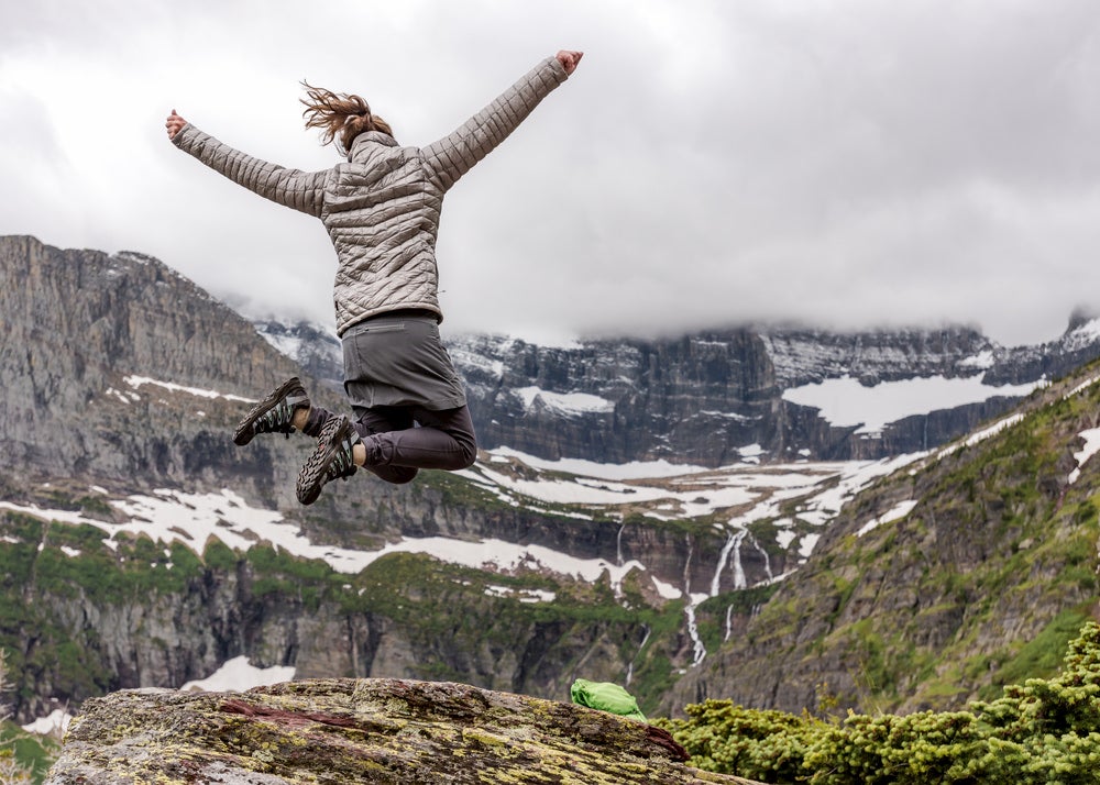 Woman in winter hiking gear jumps mid-air in front of snowy mountain range