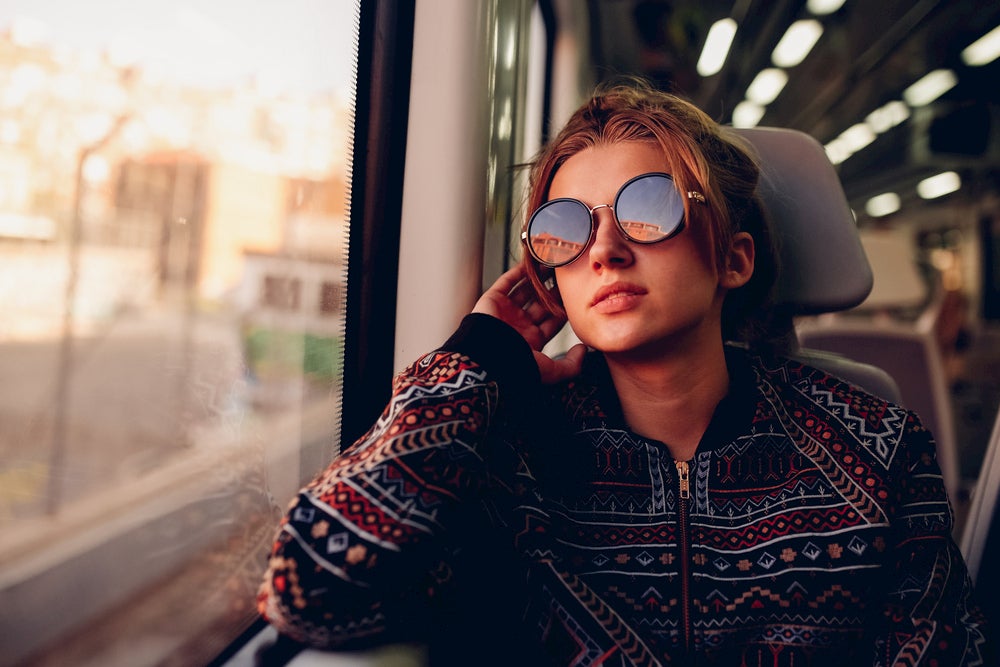 Woman wearing large, round sunglasses looks out window of train car while on the phone