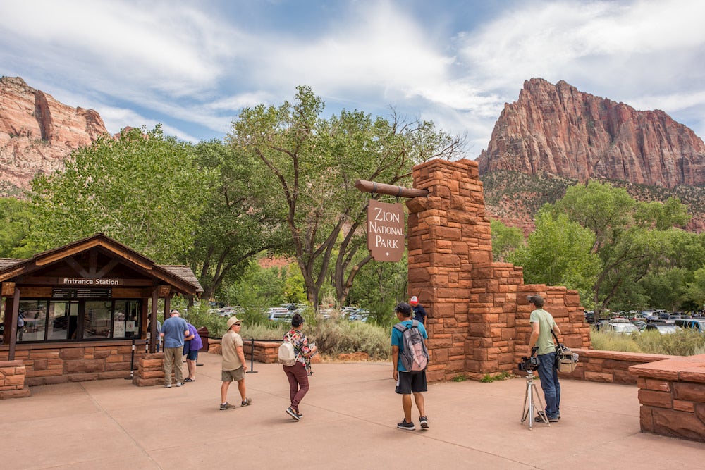 a group of visitors walk toward the entrance of Zion National Park beneath large red rock formations