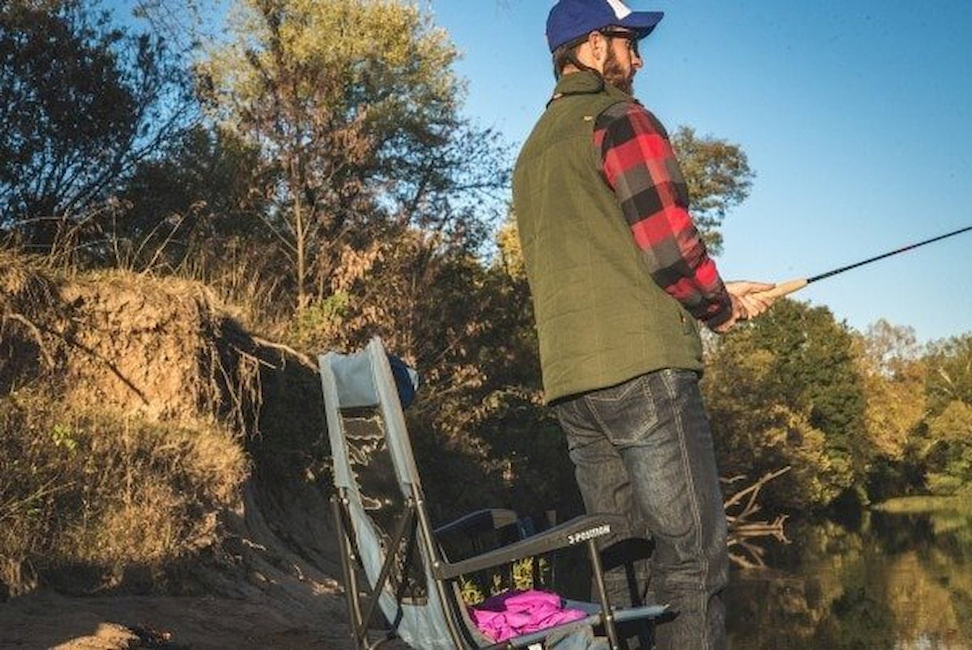 Man fishing in front of camp chair on side of calm river. 