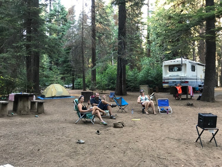 tres personas sentadas en sillas de jardín con una tienda de campaña y RV en la parte trasera en un camping boscoso