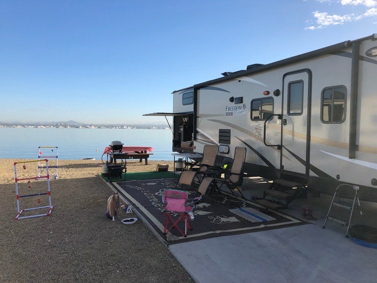 a parked RV with a rug, chairs, bathroom and games set up on a campground near a lake