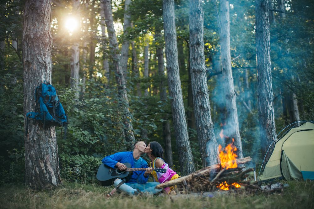 man holding guitar kisses woman beside a campfire in wooded campsite