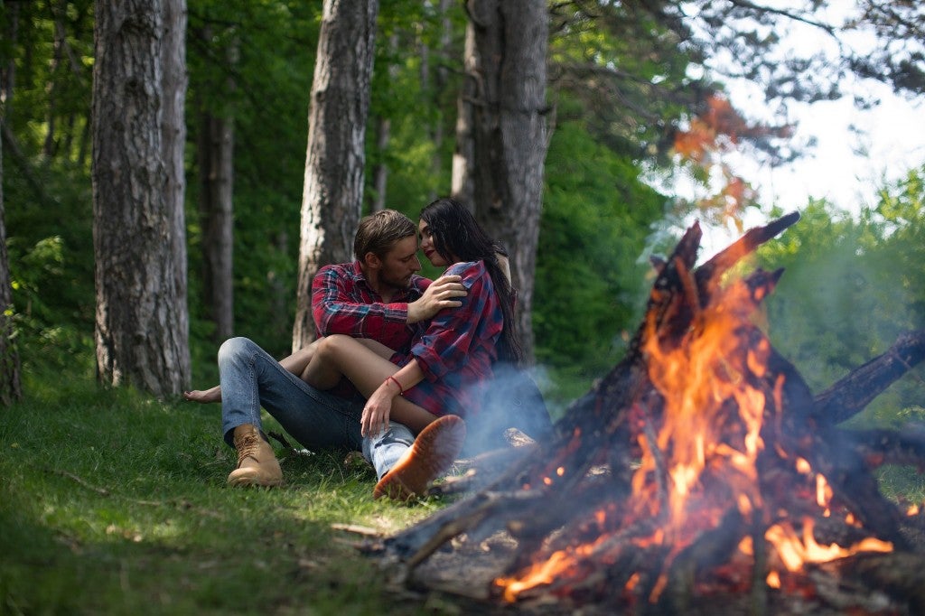 man and woman embracing in flannel shirts in front of campfire