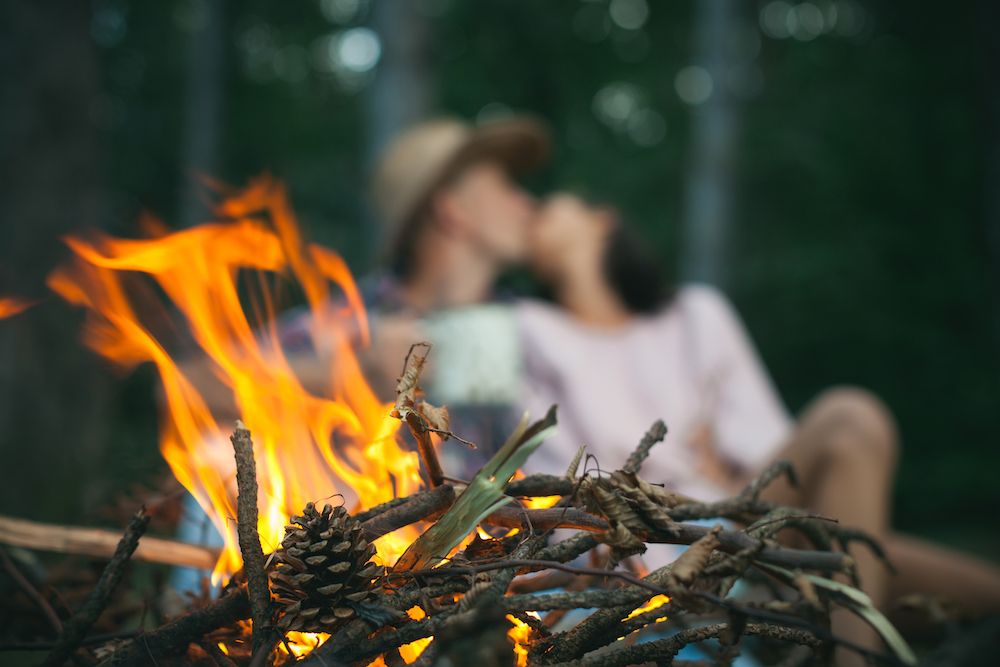 camera focuses on twigs burning in campfire as couple kisses in the background out of focus