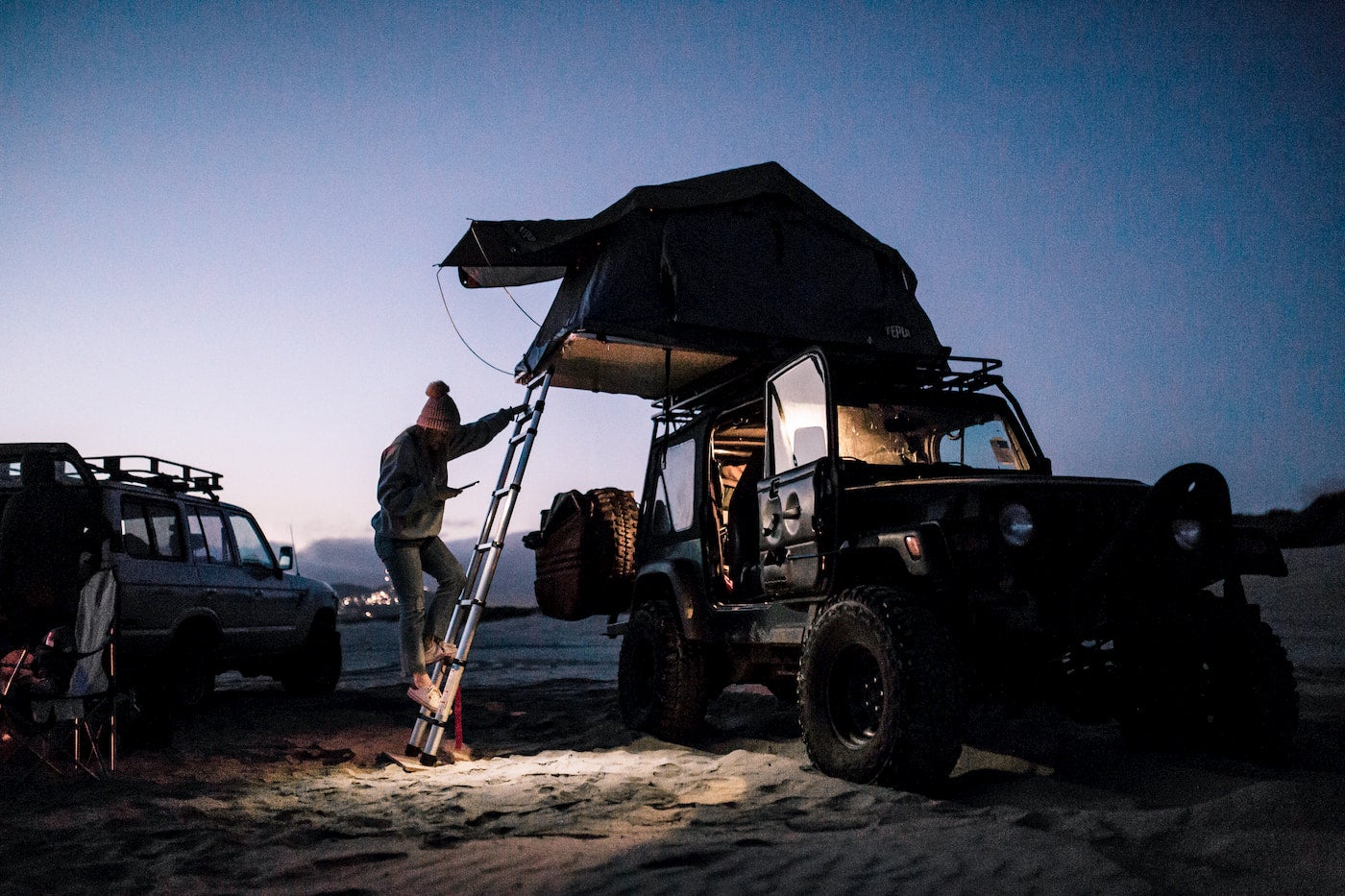 Man walking up ladder to camping car rooftop tent on the beach after dusk.