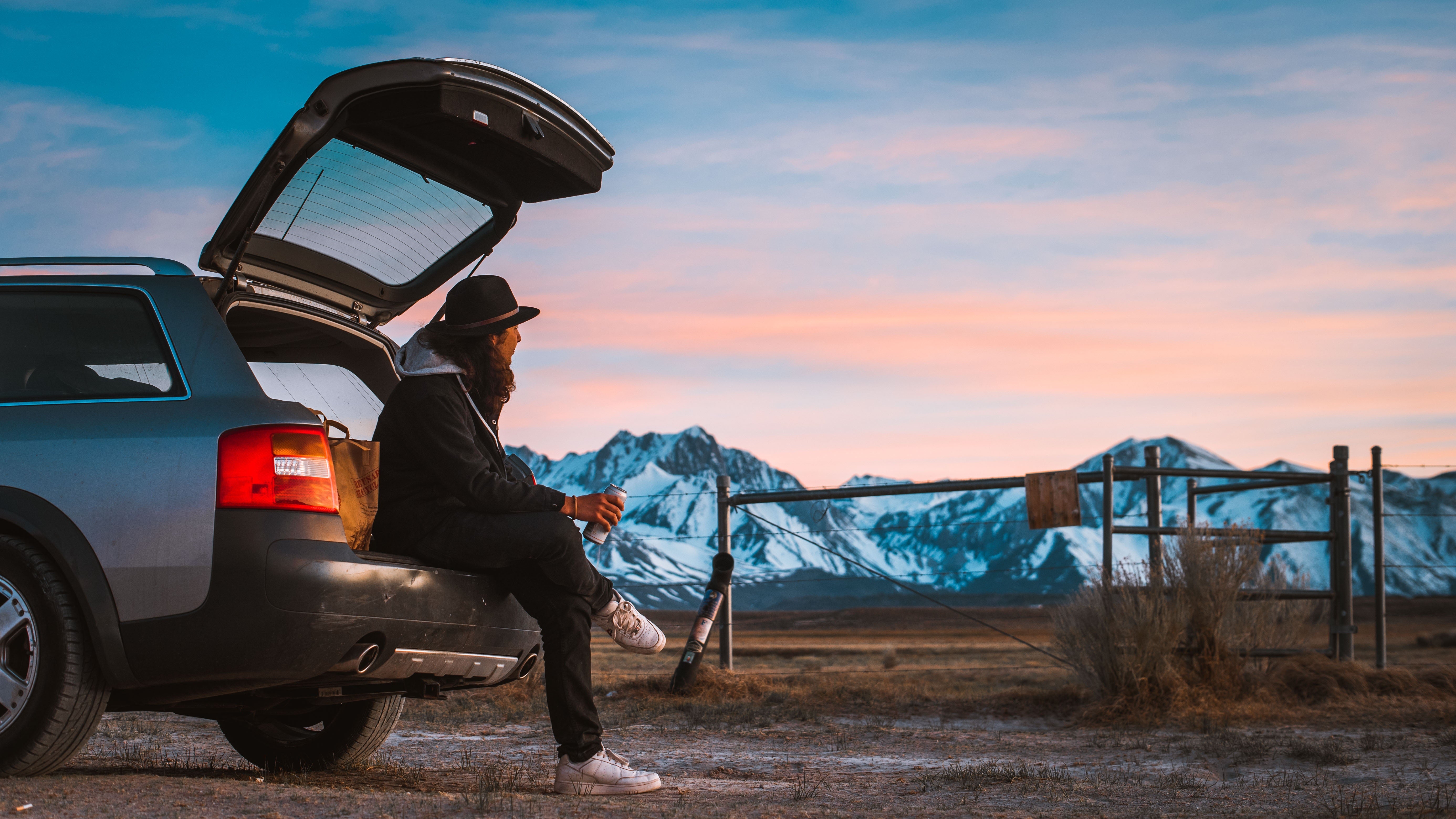 man rests on back bumper of his car with a beer while watching alpine sunset